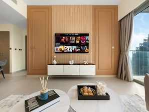 A modern living room with a flat-screen TV mounted on a wooden panel wall, a coffee table with snacks, and a view of a cityscape through large windows. - New 1BR | Burj Khalifa Infinity Pool | Dubai Mall (Dubai)