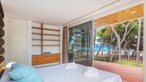 A modern bedroom with a bed, a wooden dresser, a glass door leading to a balcony with a view of the sea. - Villa Ocean - Beachfront Villa, Pool & Sea Views (Port d'Alcudia)