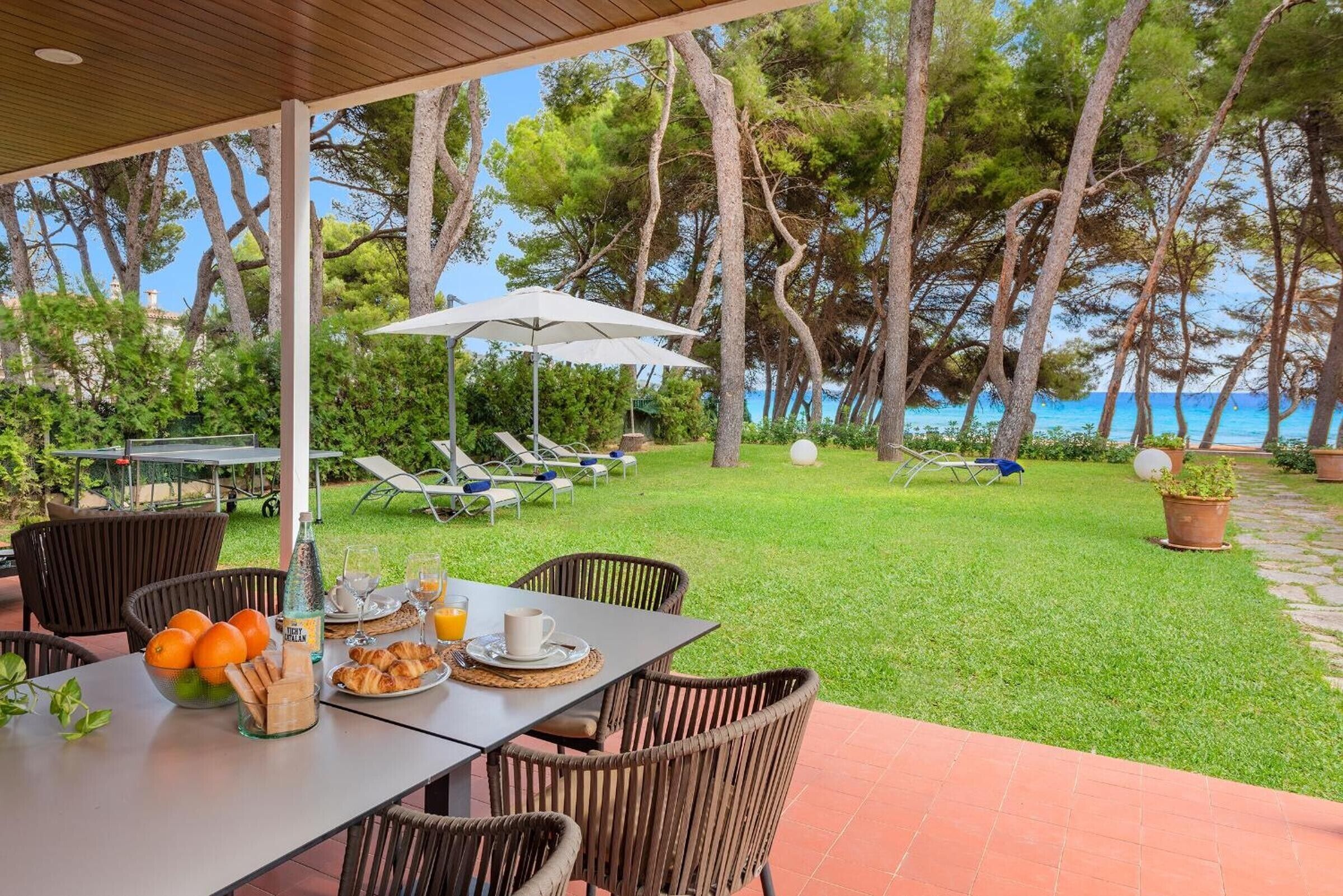 Un espace extérieur abrité, aménagé pour les repas, avec des meubles en osier, une table dressée avec des croissants, des oranges et du jus, et une vue sur la mer et les arbres.