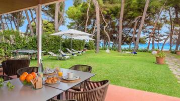 Un espace extérieur abrité, aménagé pour les repas, avec des meubles en osier, une table dressée avec des croissants, des oranges et du jus, et une vue sur la mer et les arbres.