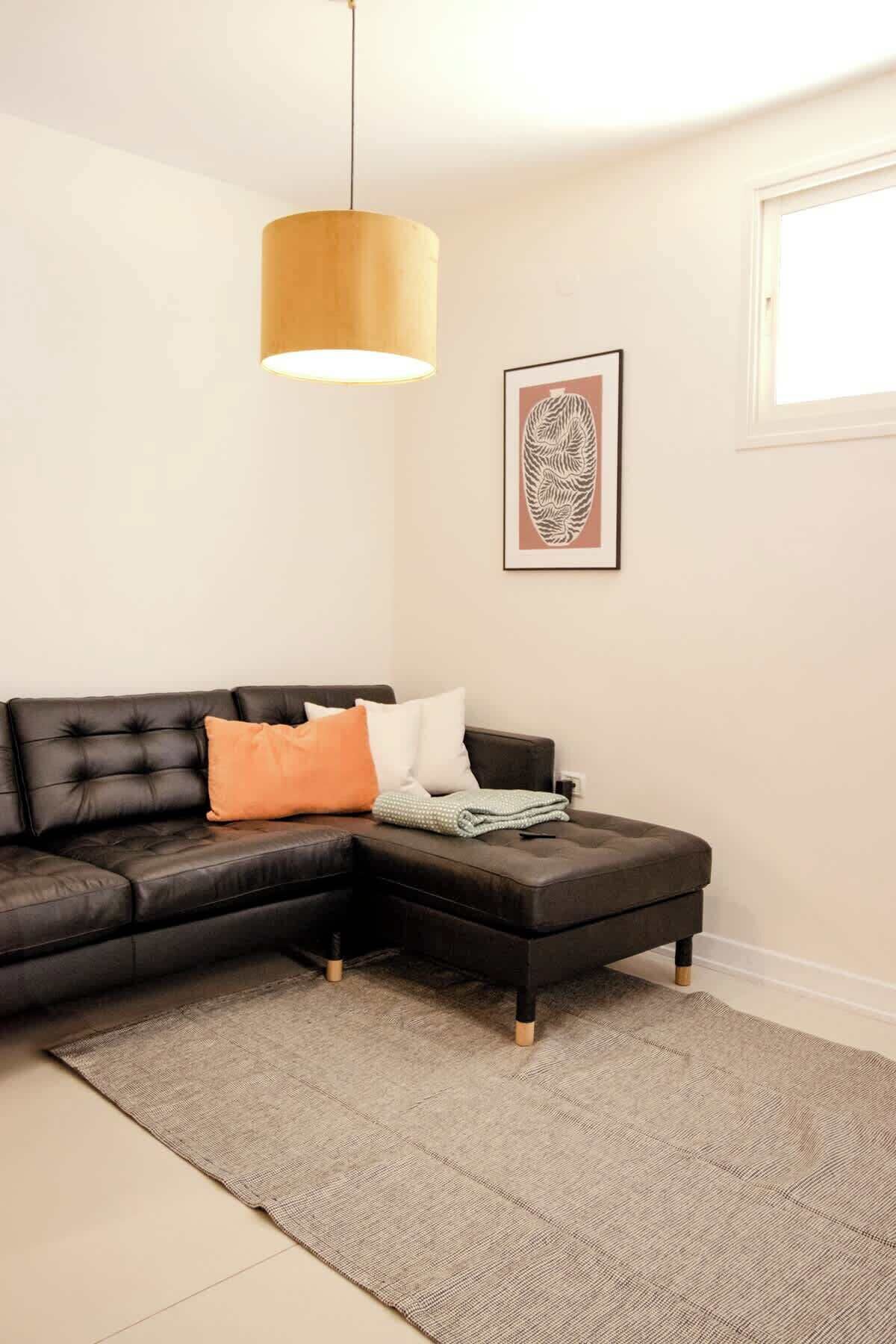 Een moderne woonkamer met een zwarte leren zithoekbank, een grijze vloerkleed, een houten salontafel, een hanglamp en een lijstwerk met een kunstwerk aan de muur.