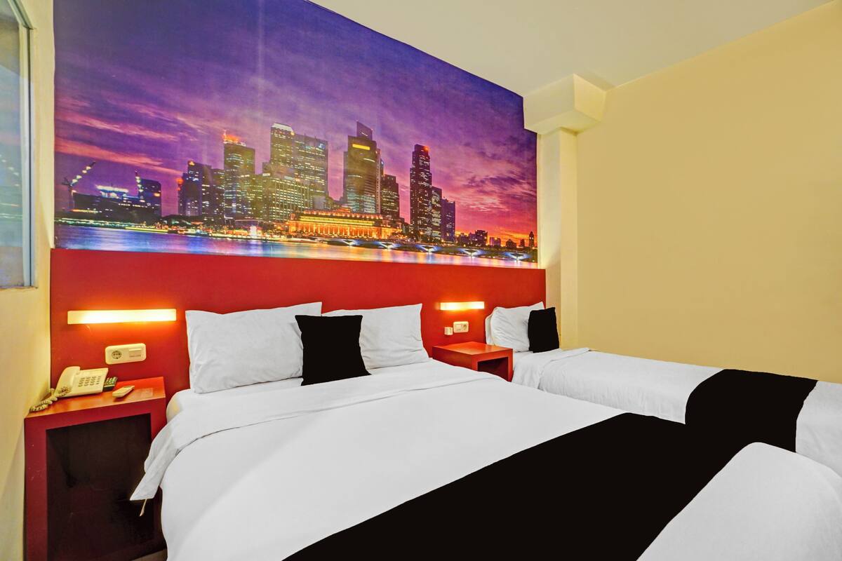 Ein Hotelzimmer mit einem großen Bett, zwei Nachttischen, einem Beistelltischchen mit Telefon und einem Wandgemälde mit Stadtansicht.