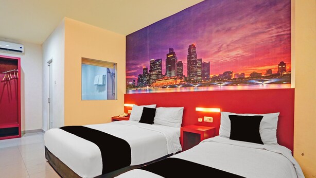Habitación de hotel con dos camas, un cuadro de un paisaje urbano en la pared, un armario rojo y una ventana con vistas a la ciudad.