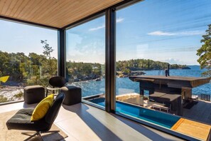 A modern room with a large window overlooking a pool and a scenic view of a rocky coastline.
