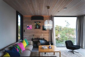 A modern living room with a wooden ceiling, a sofa with colorful cushions, a black chair, a wooden coffee table, and a view of a river and trees through large windows.