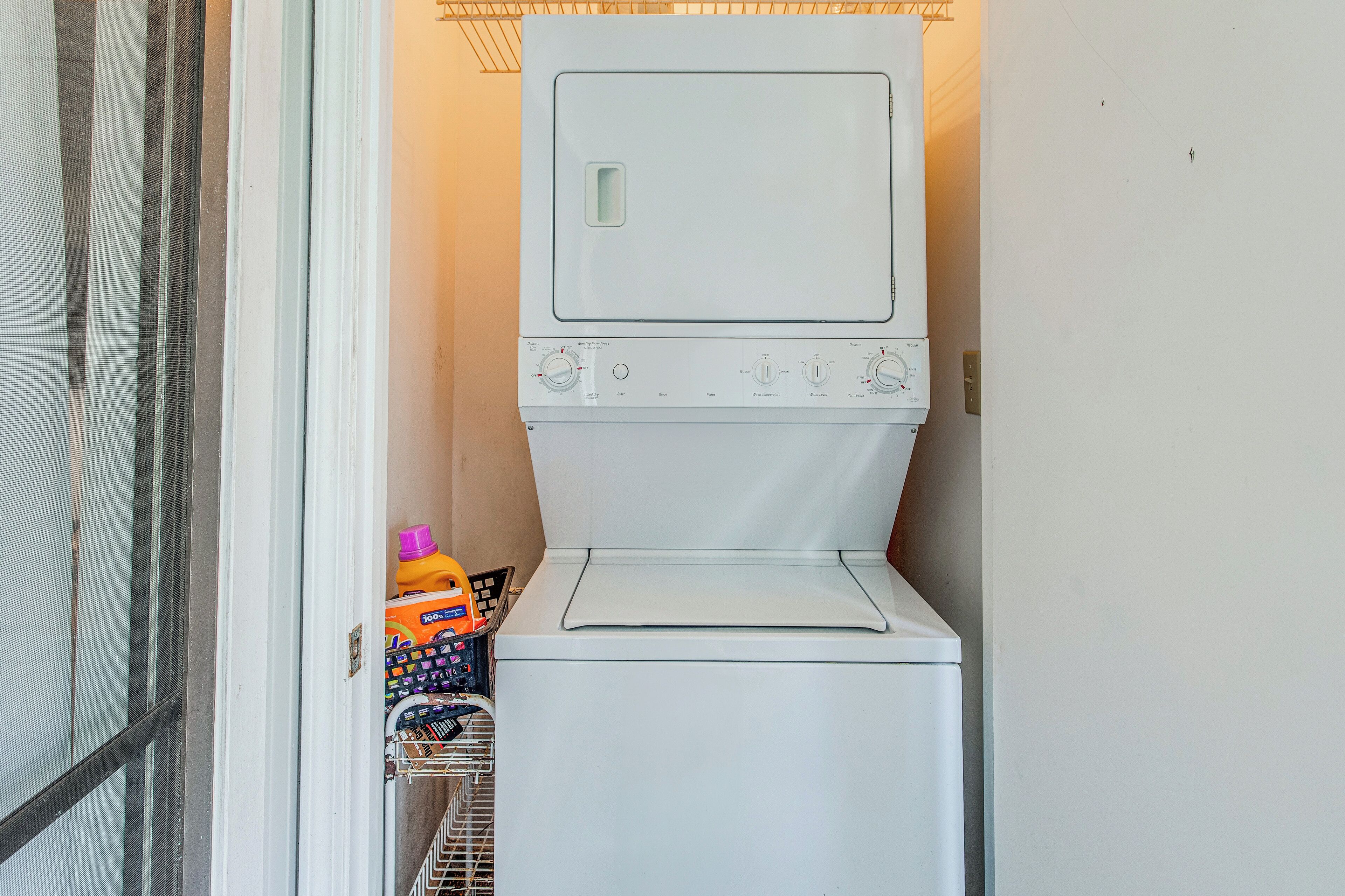 A compact, white washing machine with a front-loading door, placed in a small laundry room with a metal rack holding laundry supplies.
