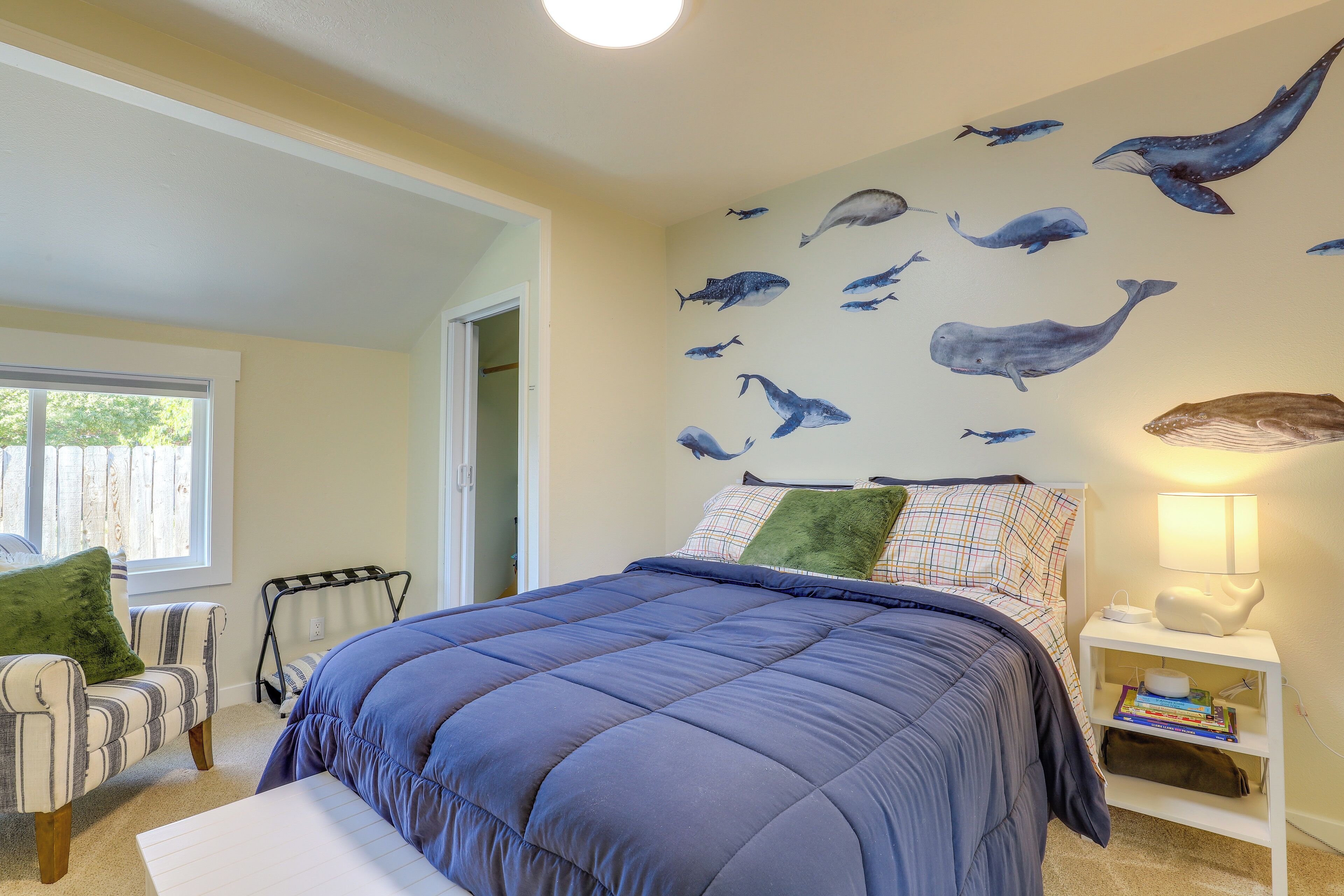 A bedroom with a large bed, a window, a chair, and a wall with whale decals.