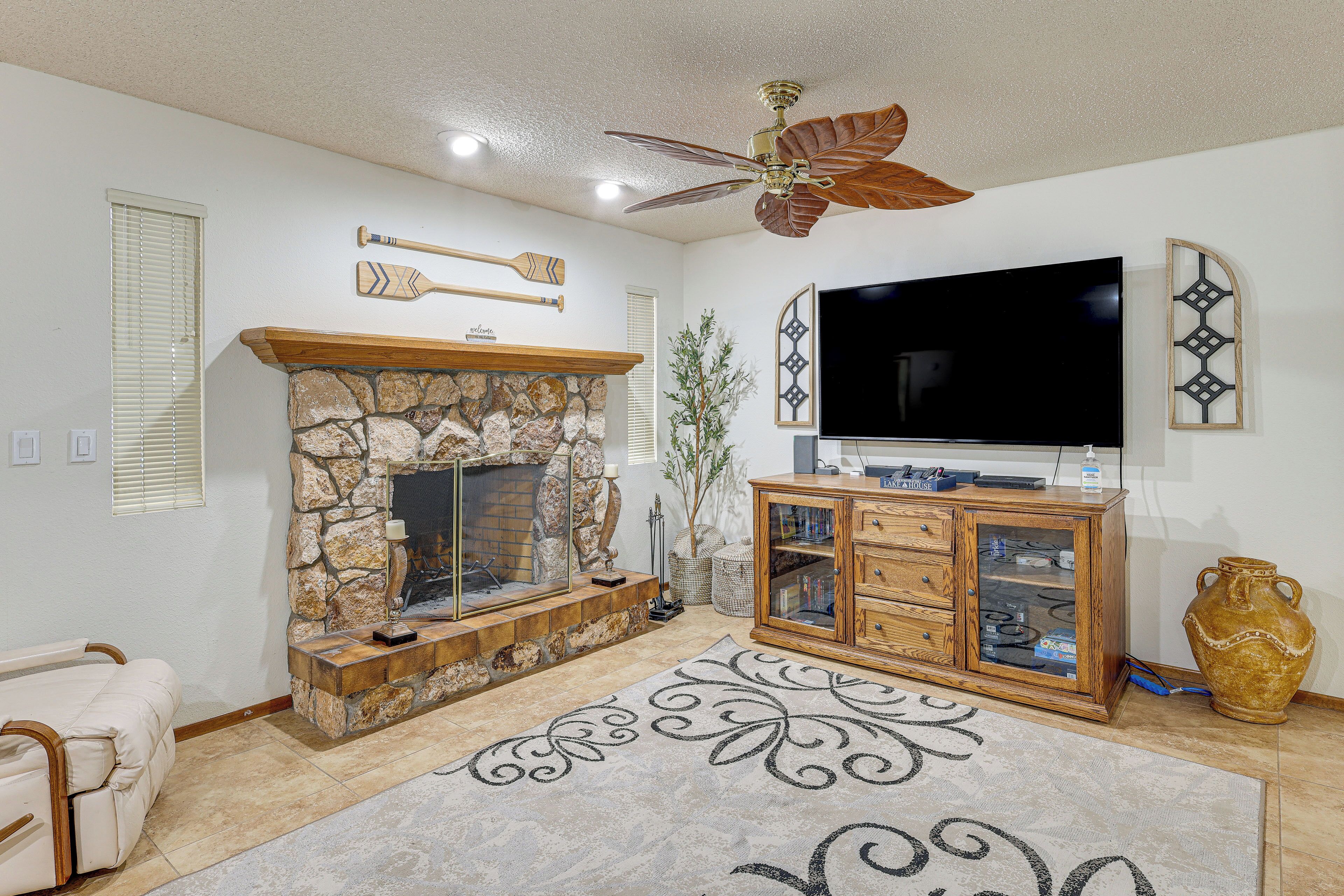 A living room with a stone fireplace, a television mounted on the wall, a wooden cabinet, and a ceiling fan.