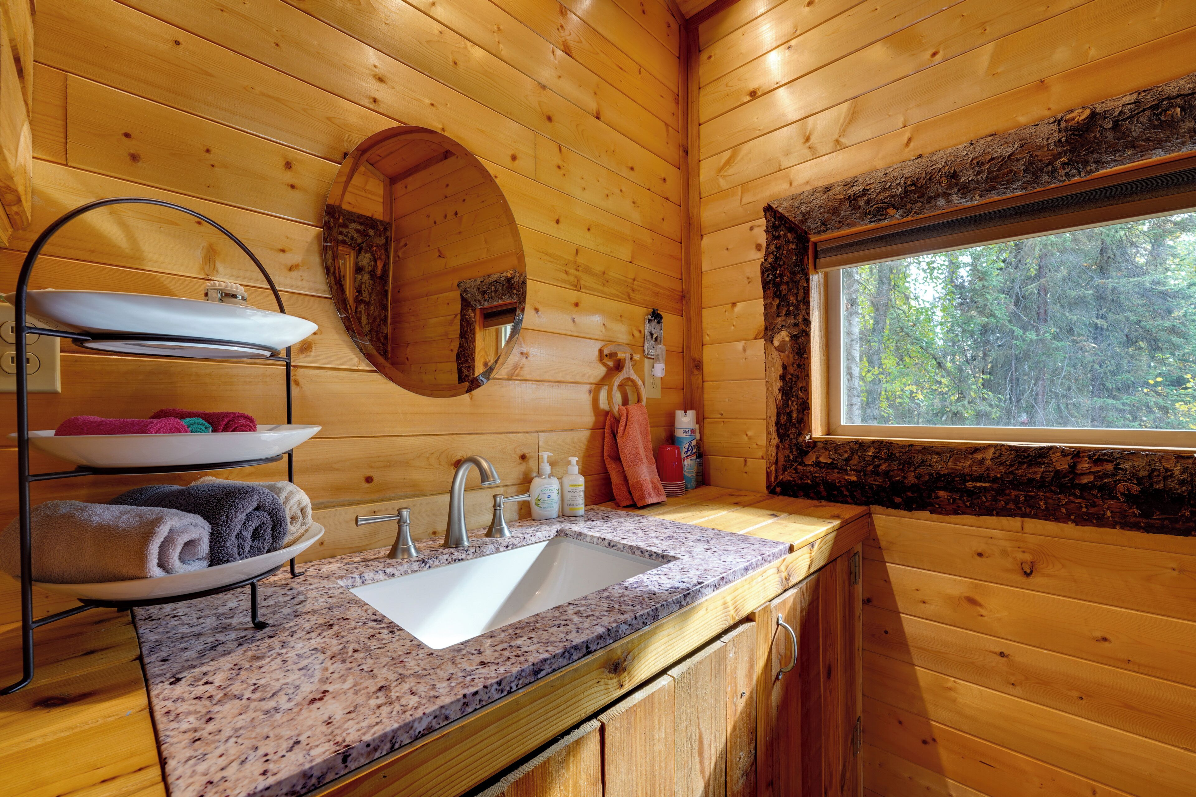 Baño de cabaña de madera con lavabo de granito, espejo redondo y ventana con vista a árboles.