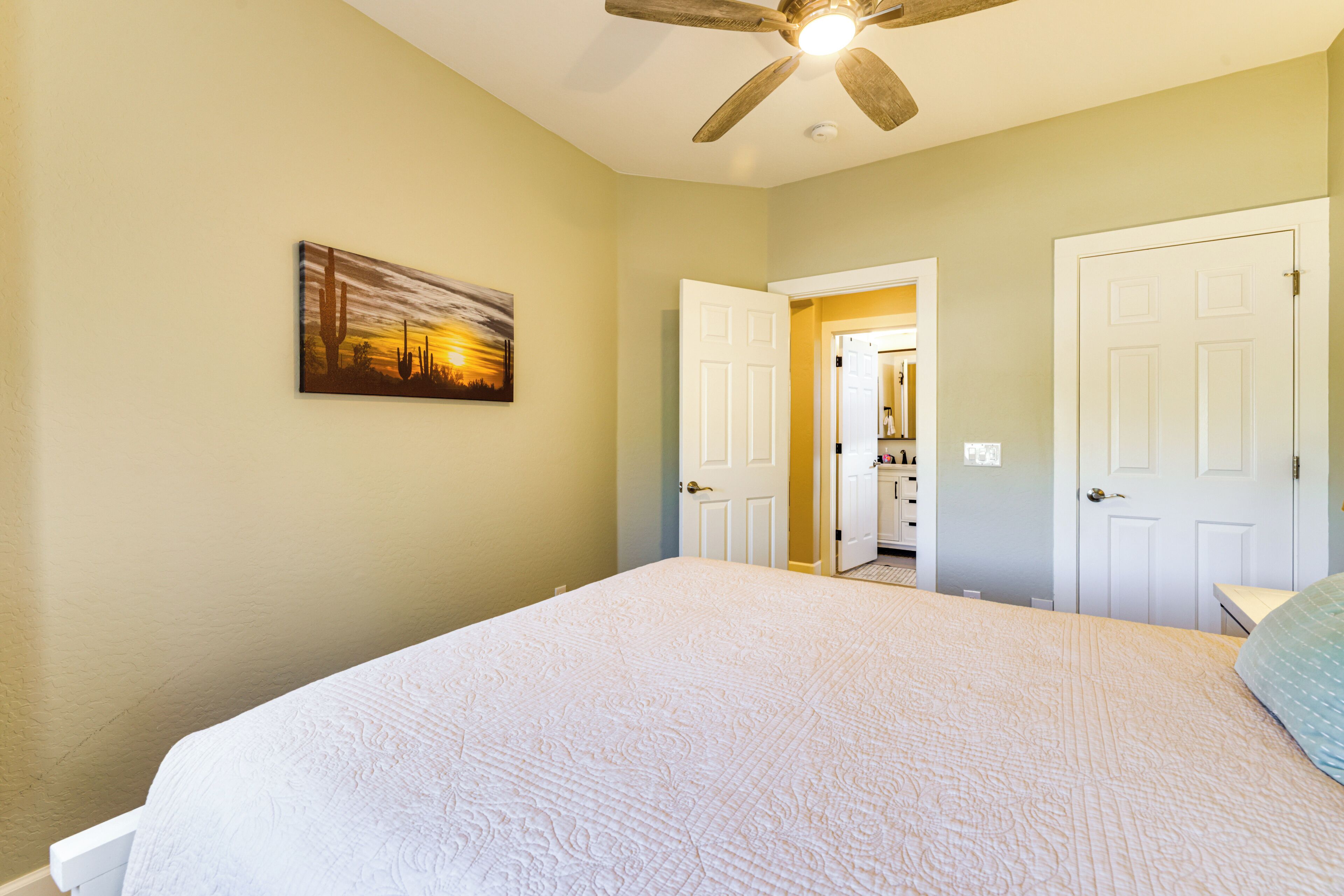 A bedroom with a bed, a ceiling fan, a door, and a painting on the wall.