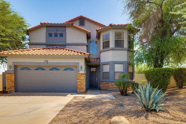 A two-story house with a garage, a front door, and a small garden with a plant. - Pool, Putting Green, West Valley Family Home (Goodyear)