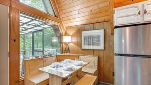 A wooden cabin interior with a dining table set for four, a bench, a refrigerator, and a large window overlooking a deck.