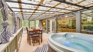 A covered outdoor patio with a hot tub, wooden furniture, and glass doors leading to a garden.