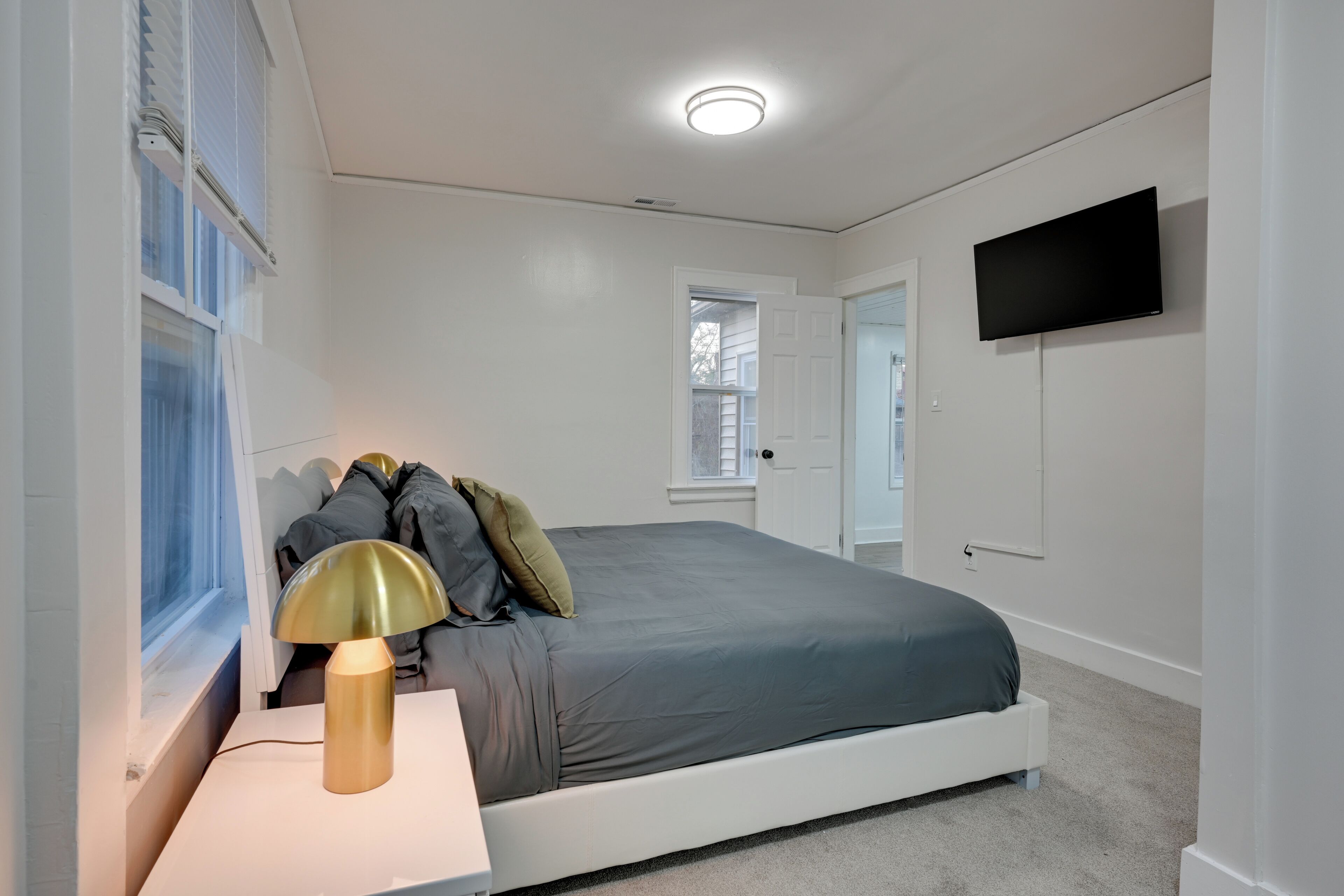A bedroom with a bed, a bedside table with a lamp, a TV on the wall, and a window with blinds.