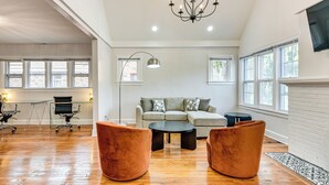 A spacious living room with hardwood flooring, a grey sofa set, two brown armchairs, a black coffee table, and a chandelier.