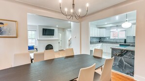 A modern dining area with a dark wooden table and white chairs, a chandelier, and a kitchen visible through an open doorway.