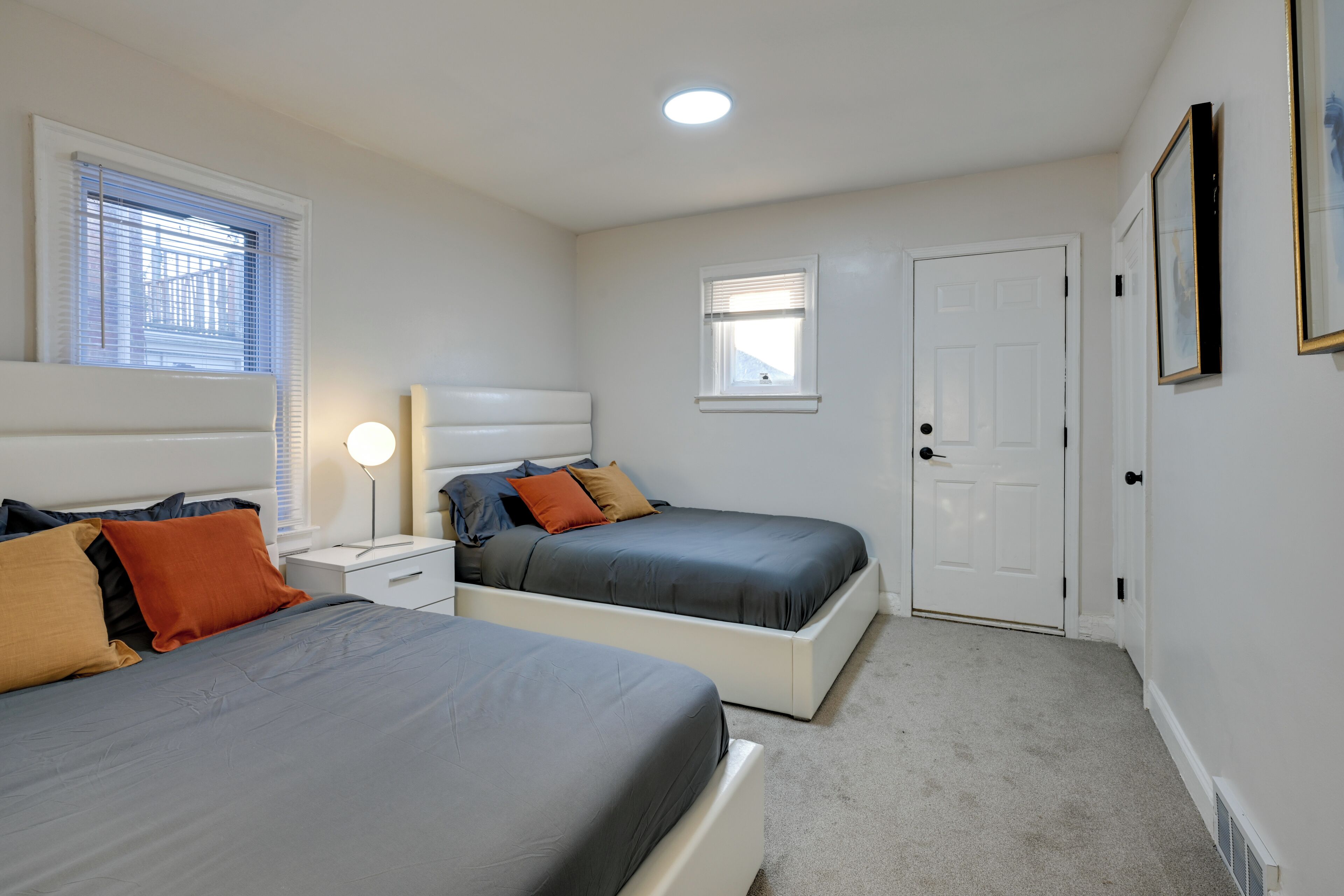 A bedroom with two beds, a nightstand, a bedside table with a lamp, a window with blinds, and a door.
