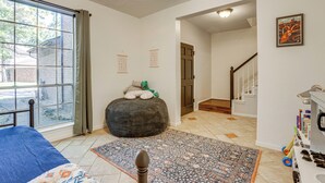 A bedroom with a large window, a bed, a round ottoman, a patterned rug, and a staircase leading to another room. - Bike & Explore: Family Home in The Woodlands! (The Woodlands)