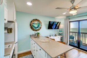 A kitchen with a large island, a ceiling fan, a TV mounted on the wall, and a view of the outdoors through a sliding glass door.