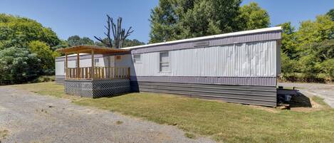 A mobile home with a covered porch and a small deck, surrounded by a gravel driveway and grassy area.