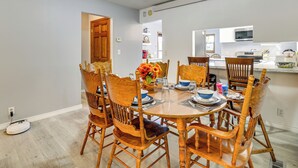 A dining area with a round wooden table set for four, surrounded by matching chairs. A vase with flowers is on the table, and there is a kitchen visible in the background.