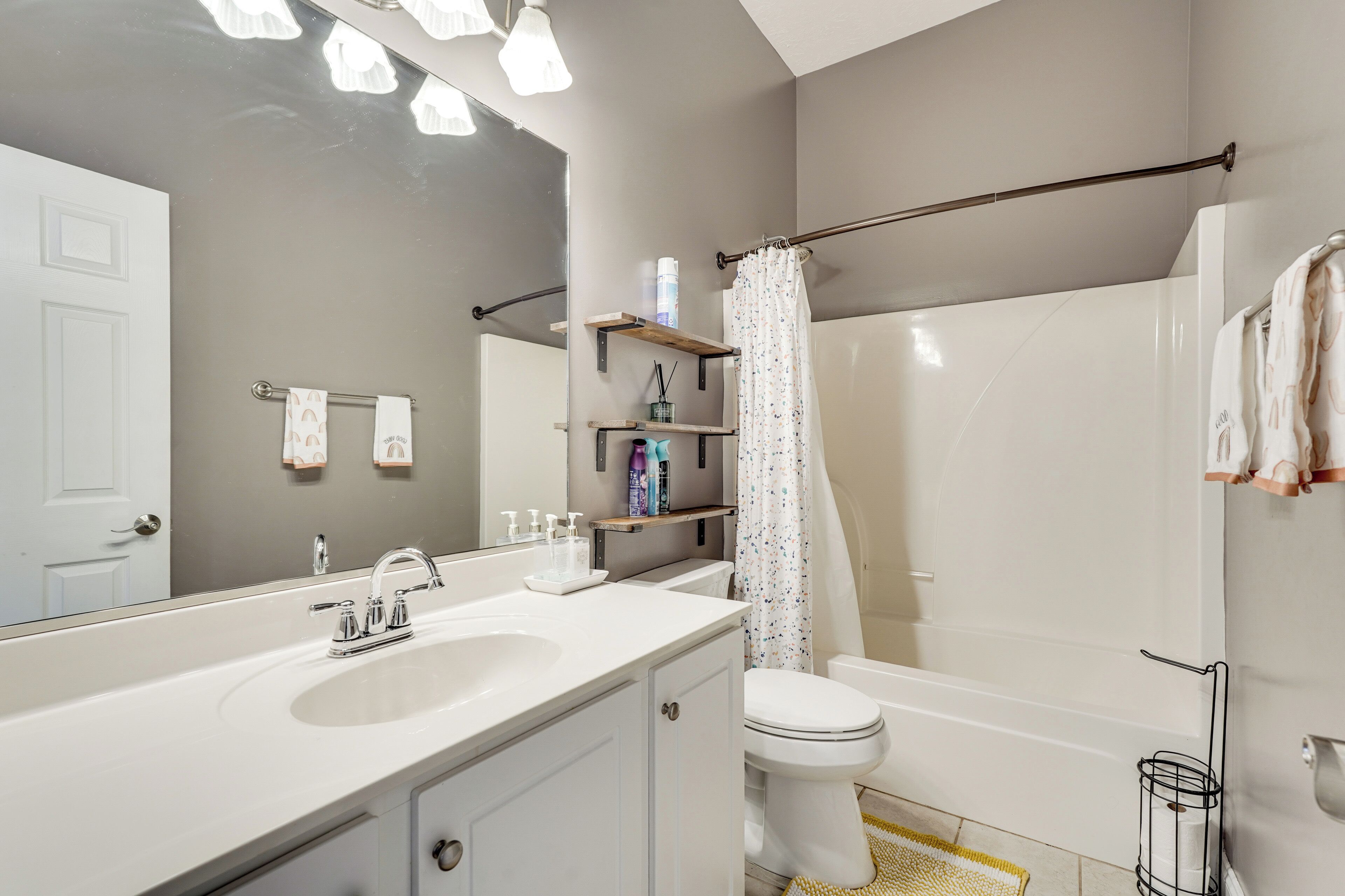 A modern bathroom with a white sink, mirror, and toilet. There are shelves with toiletries and a shower curtain.