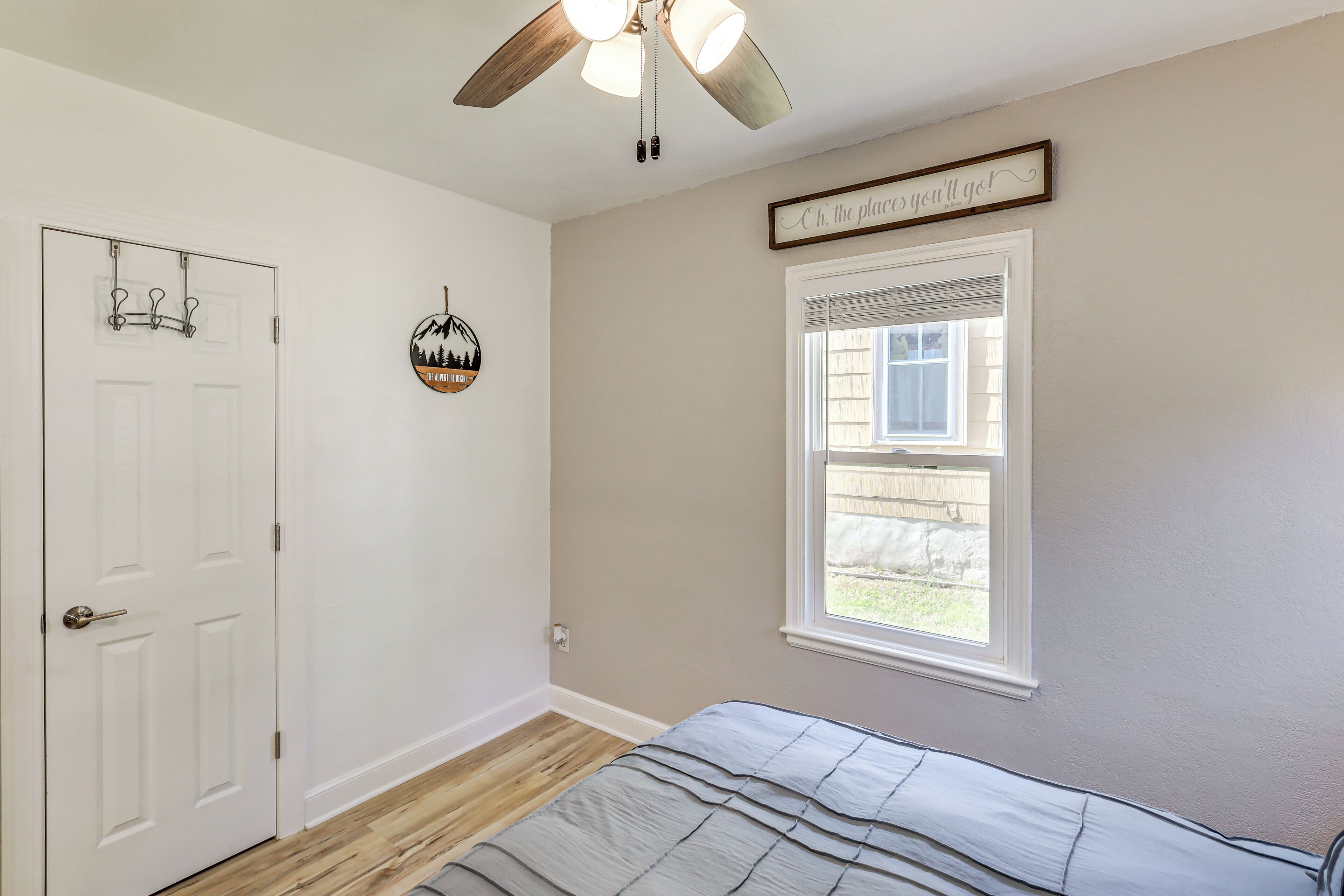 Une chambre à coucher comprenant un lit, un ventilateur de plafond, une fenêtre avec des stores, une porte avec un porte-manteau et un élément décoratif mural.