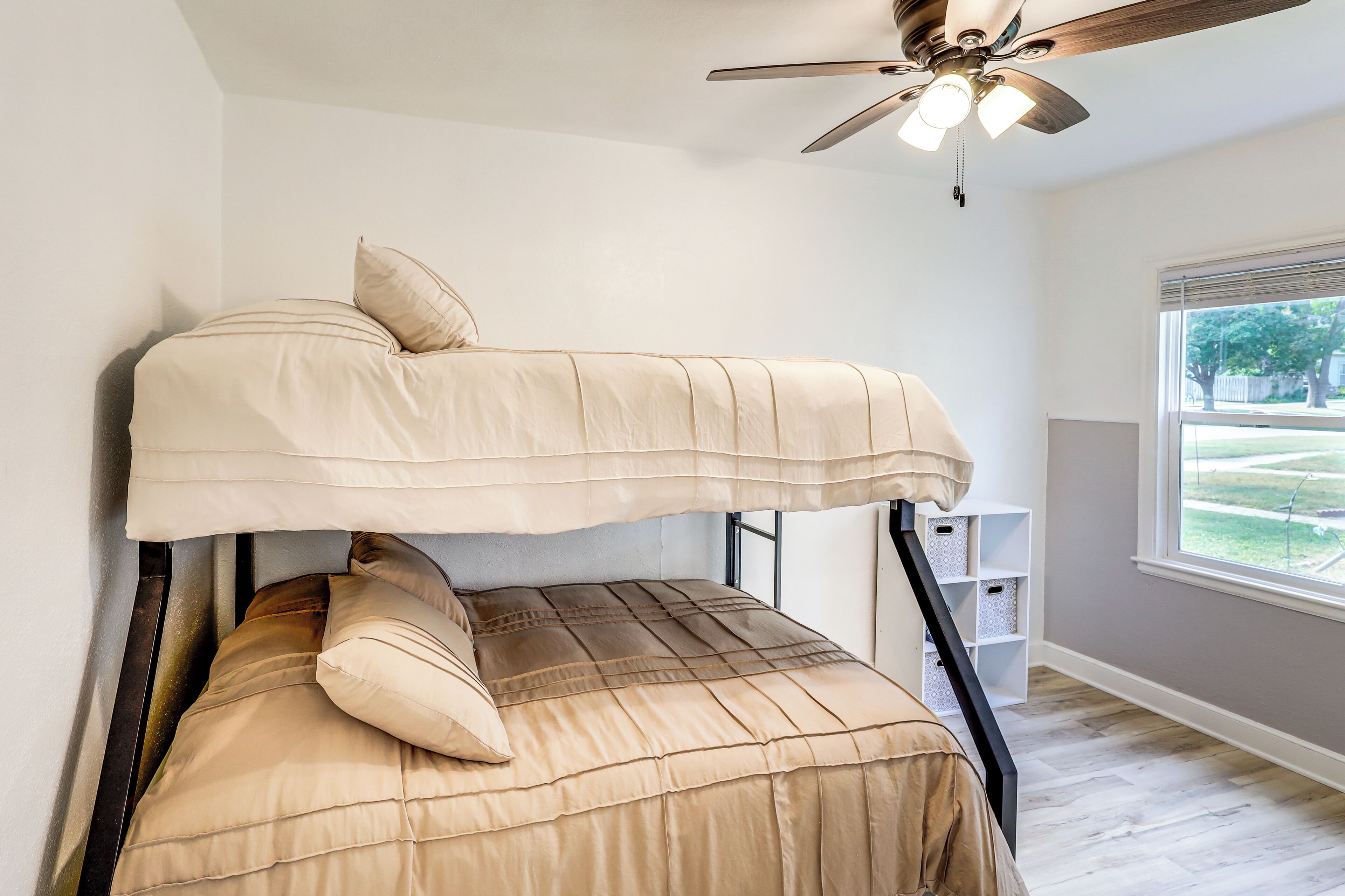 A bunk bed with beige bedding, a ceiling fan, and a window with a view of a yard.