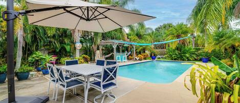 A poolside area with a table and chairs, a covered seating area, and a pool.