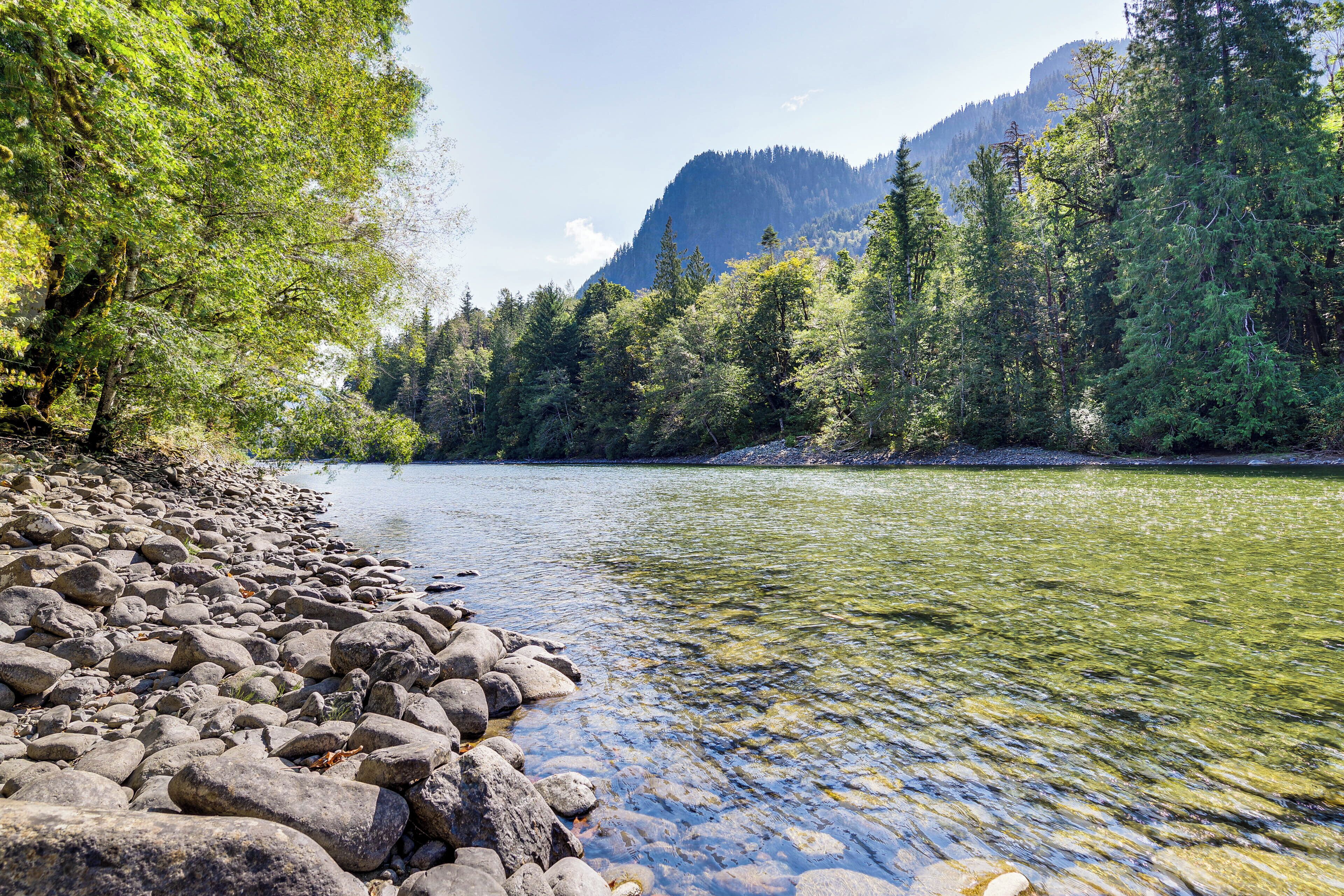 A riverbank with clear water, surrounded by trees and mountains.