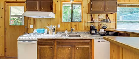 A kitchen with wooden cabinets, a stove, a sink, and a window with a view of trees.