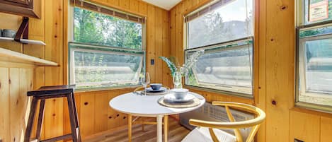 A wooden interior with a round table, two chairs, a bar stool, and a window with a view of greenery.