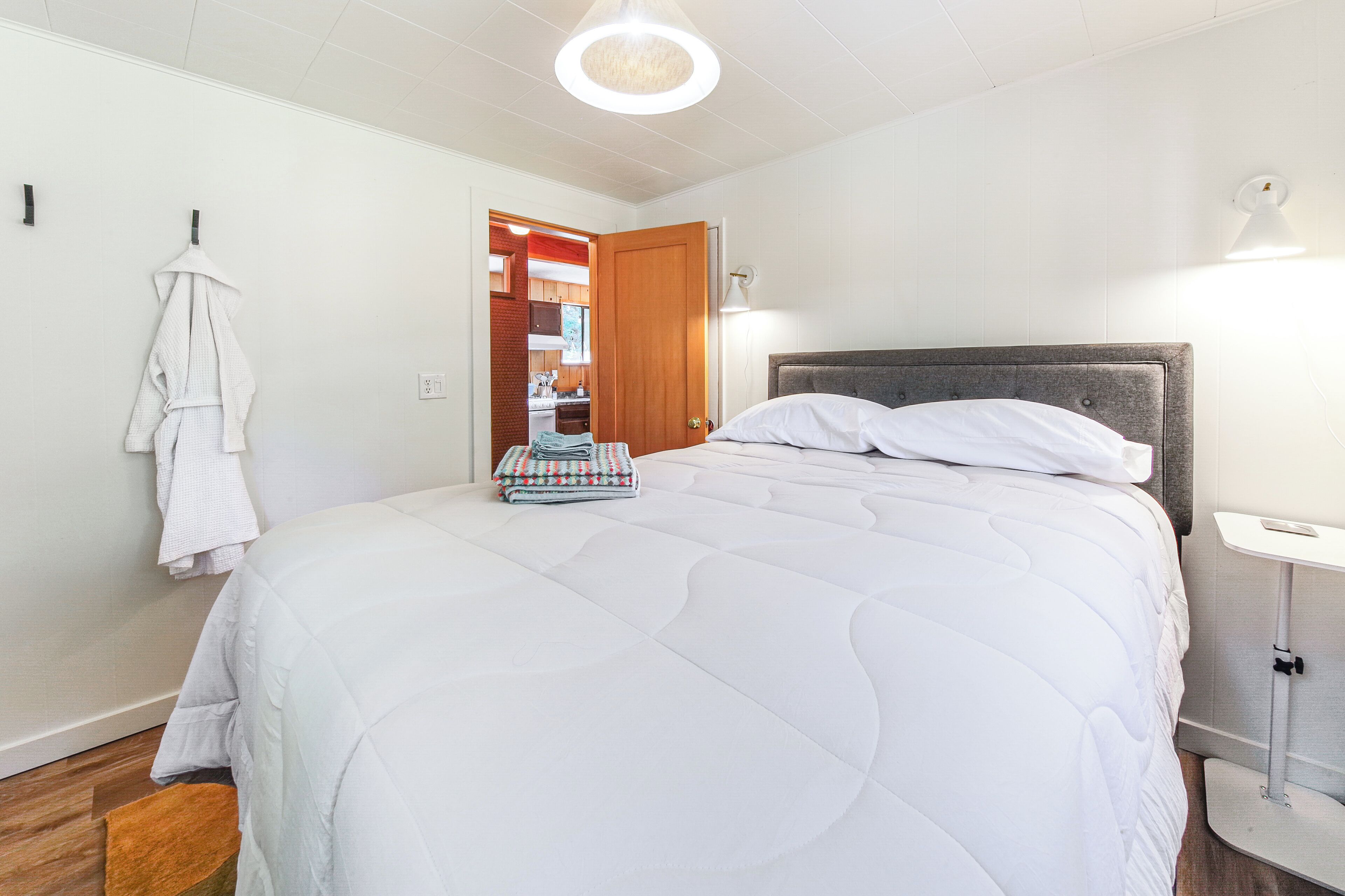 Uma cama bem arrumada com edredom branco, uma mesa de cabeceira com abajur, e vista para a área da cozinha.