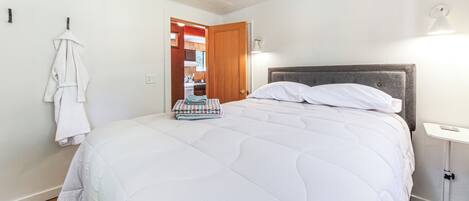 Uma cama bem arrumada com edredom branco, uma mesa de cabeceira com abajur, e vista para a área da cozinha.