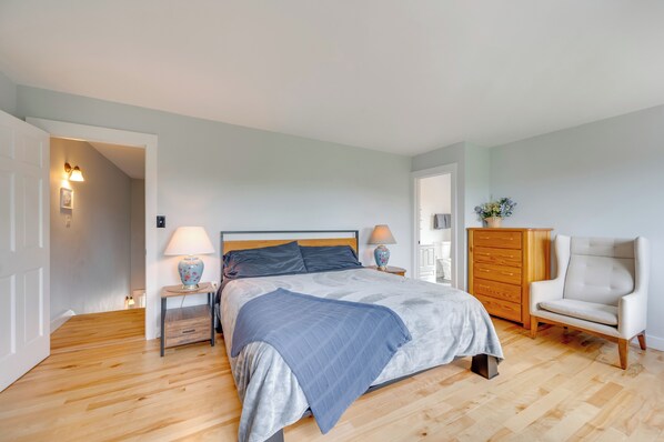 Une chambre à coucher comprenant un lit, des lampes de chevet, une commode, une chaise et une porte donnant sur une autre pièce.