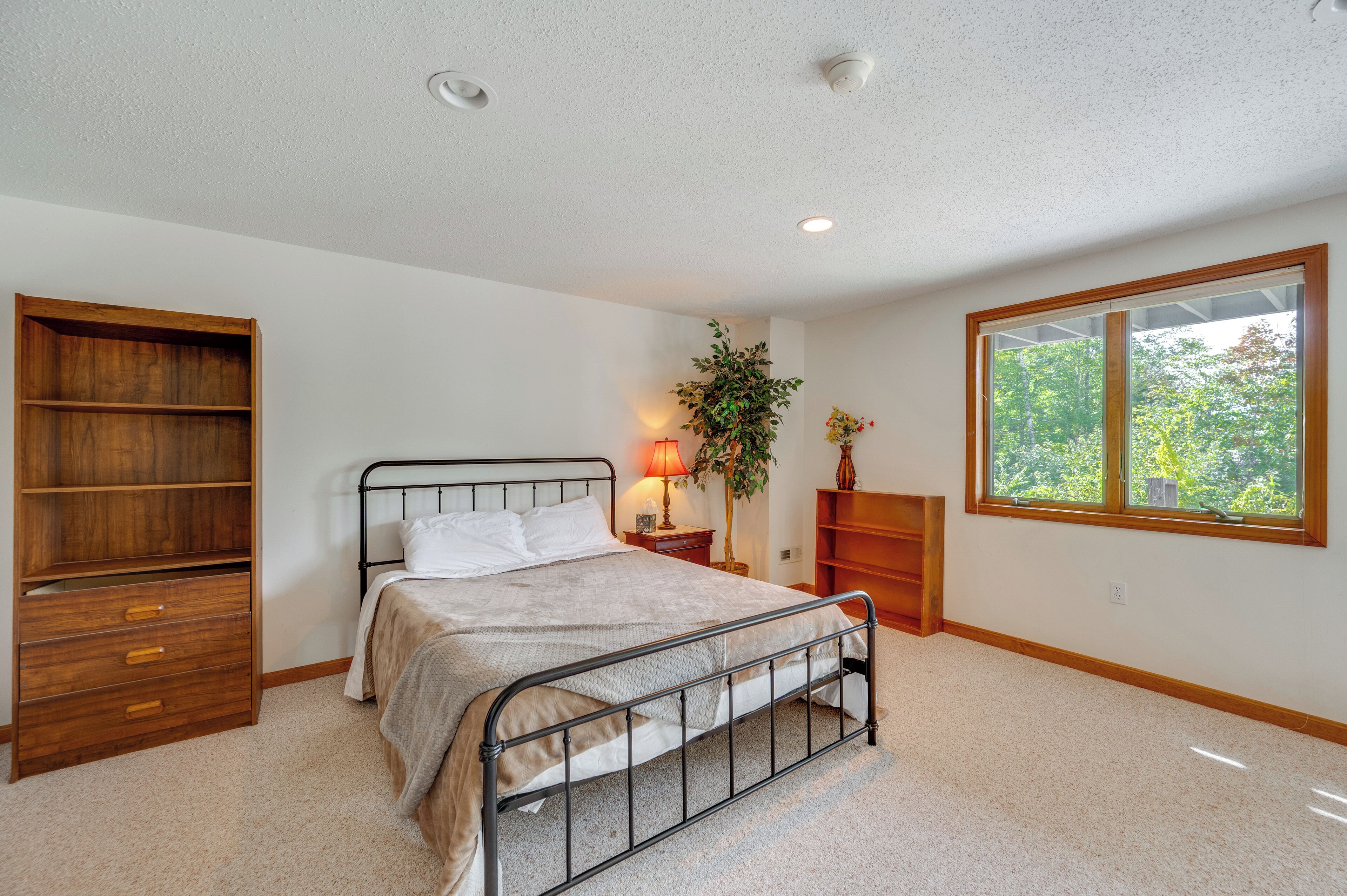 Une chambre à coucher comprenant un lit, une armoire en bois, une table de chevet avec une lampe et une fenêtre donnant sur des arbres.