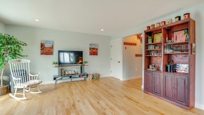 Amplia sala de estar con piso de madera, una mecedora blanca, un televisor y un mueble de madera con cristalería.