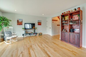 Un salon spacieux doté d’un plancher en bois, d’un fauteuil à bascule blanc, d’un téléviseur et d’un meuble en bois contenant de la verrerie.