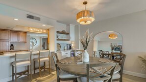 A modern kitchen with wooden cabinets, a central island, and a dining area with a round table and chairs. - Gulf Views, Steps to Beach: Redington Shores Condo (Redington Shores)