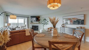 A modern living room with a wooden dining table, chairs, and a sofa. There is a fireplace, a television, and a large window. - Gulf Views, Steps to Beach: Redington Shores Condo (Redington Shores)