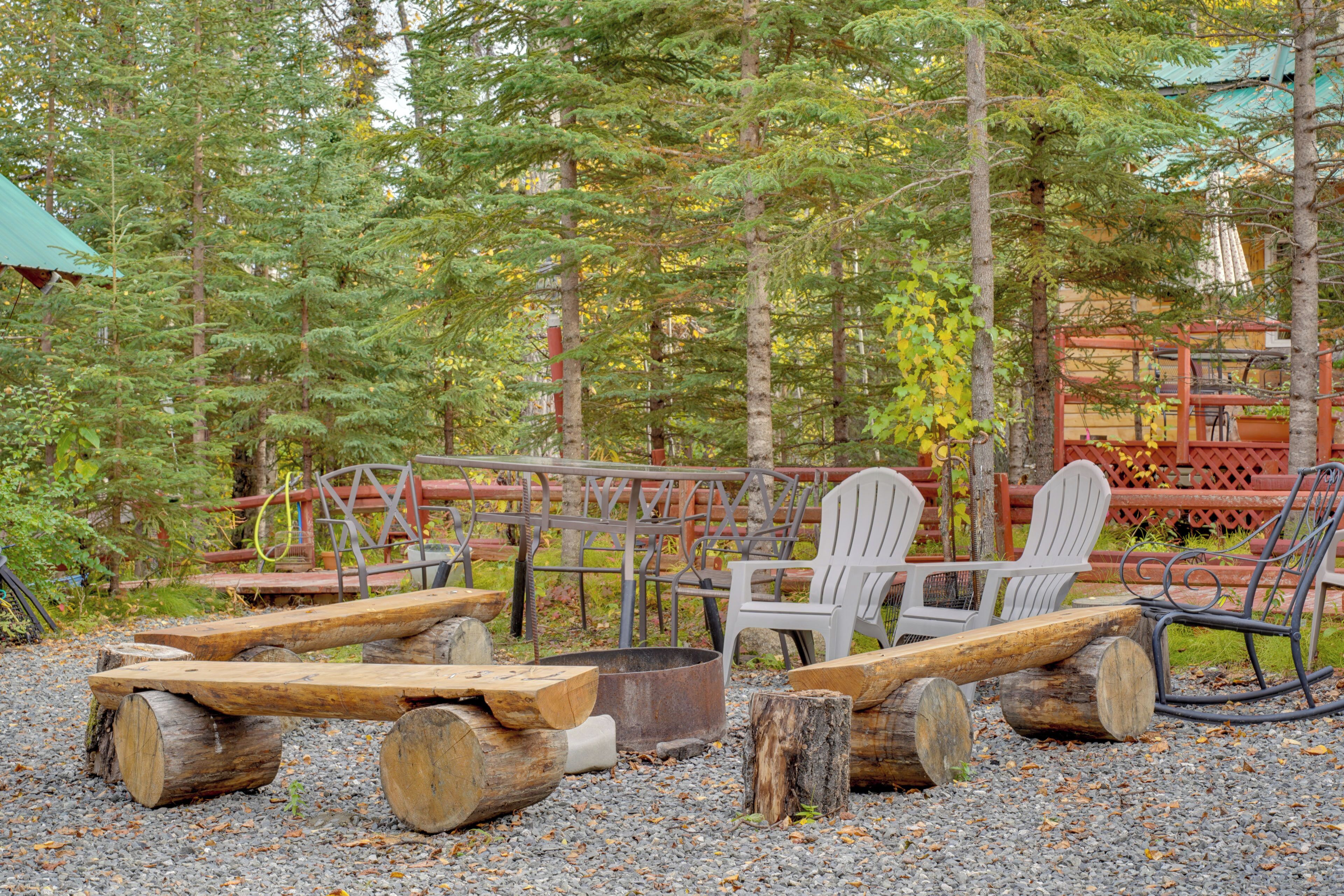 A rustic outdoor seating area with Adirondack chairs, a fire pit, and wooden logs.