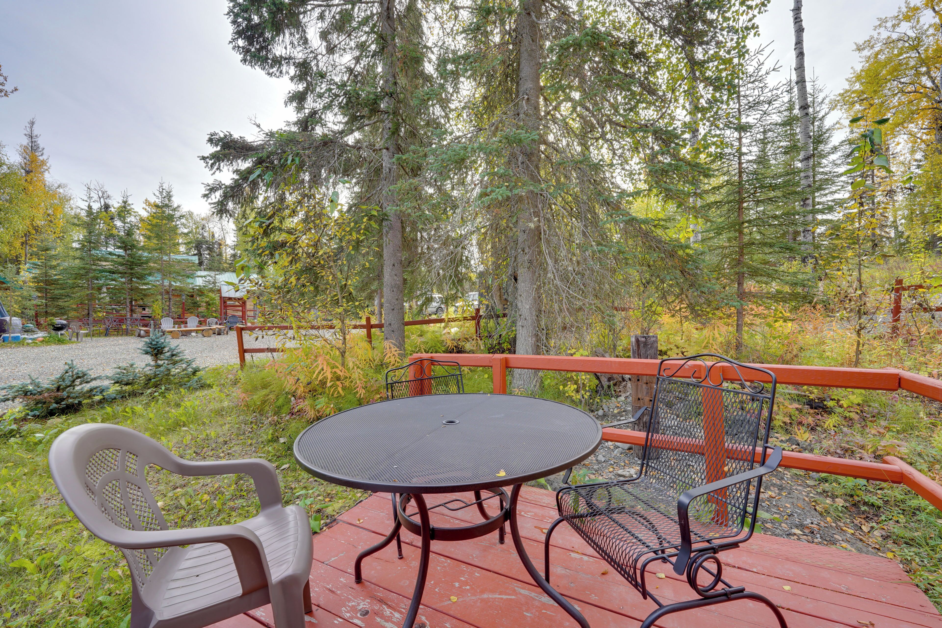 A wooden deck with a round table and two chairs, surrounded by a red railing, overlooking a forested area.