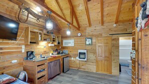 A wooden cabin interior with a kitchen area, wooden walls, a television, and a clock. - Fish, Hike & Explore: Lakefront Cabin in Soldotna (Soldotna)