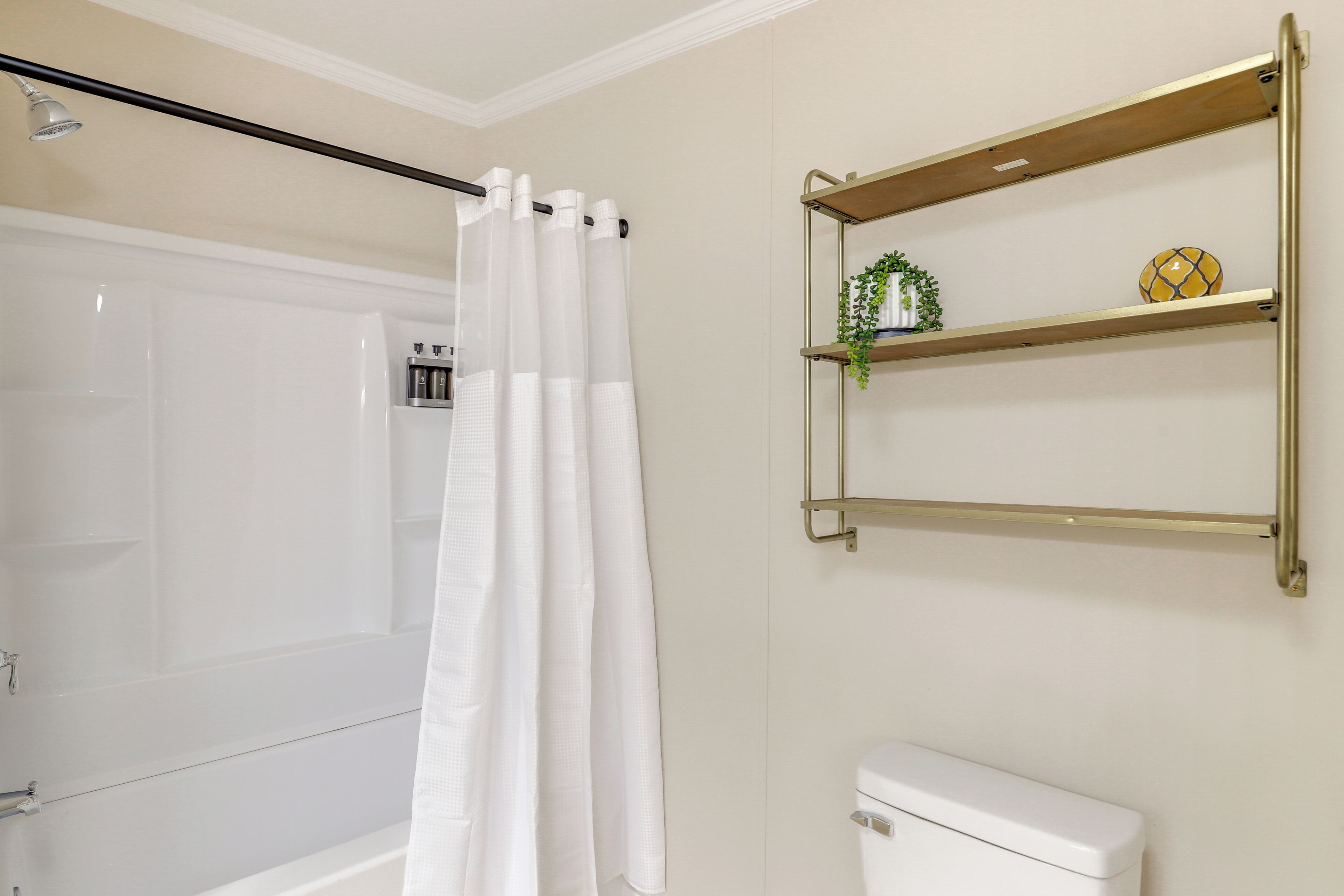 Un baño con una bañera blanca, una cortina de ducha y un inodoro. Hay dos estantes con una planta y un objeto decorativo.
