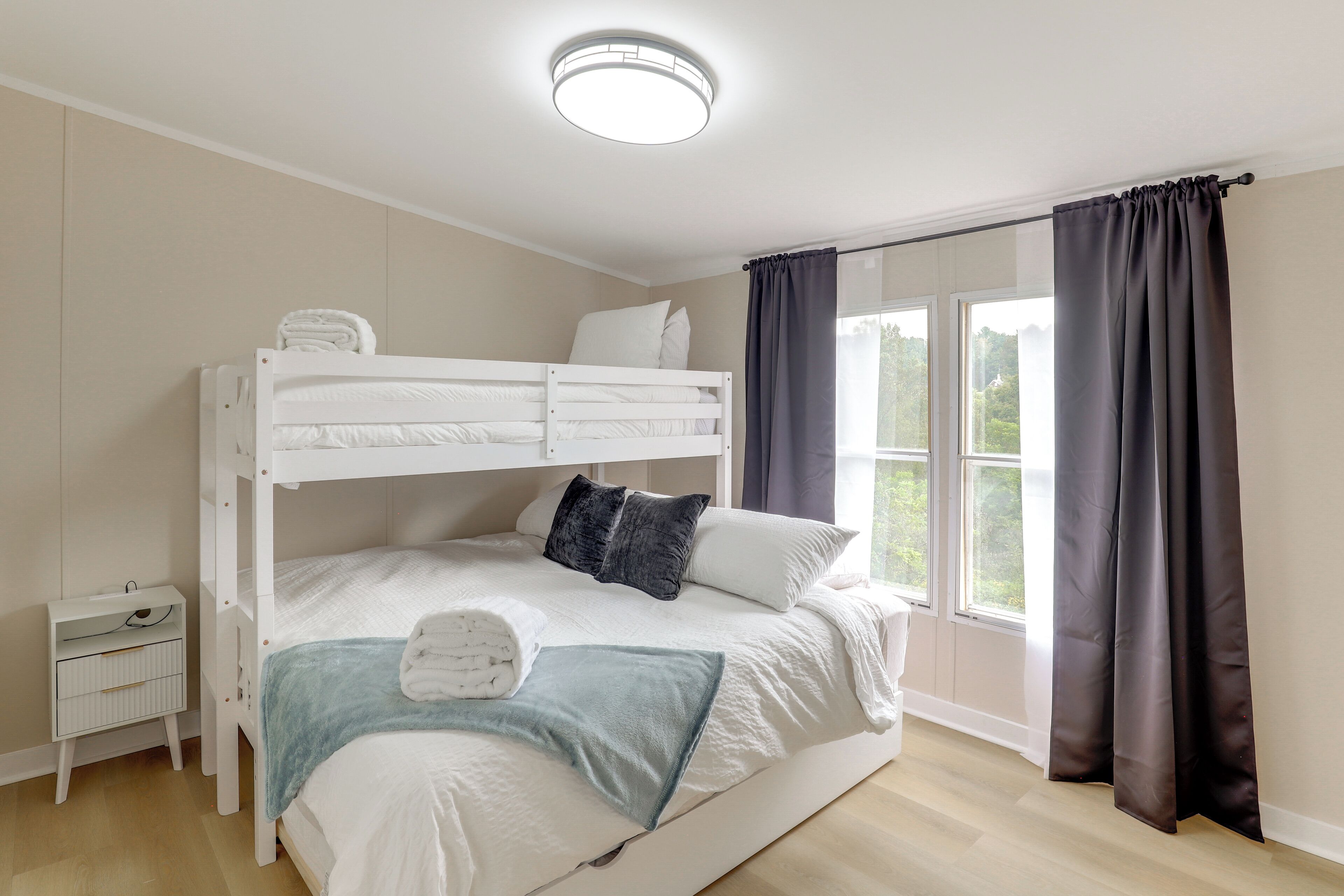 A bunk bed room with a bedside table, a window with curtains, and a ceiling light.