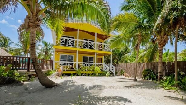A two-story yellow building with a balcony, surrounded by palm trees and a sandy ground. - Nemo's Loft: Panoramic 2BR/2BA Beachfront View in Ambergris Caye (San Pedro)