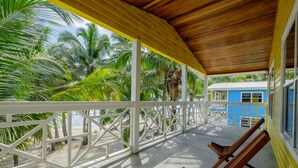A covered balcony with white railings, wooden chairs, and a view of palm trees and a blue building. - Nemo's Loft: Panoramic 2BR/2BA Beachfront View in Ambergris Caye (San Pedro)