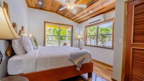 A bedroom with a wooden ceiling, a bed with white bedding, two windows with views of palm trees, and a ceiling fan. - Nemo's Loft: Panoramic 2BR/2BA Beachfront View in Ambergris Caye (San Pedro)