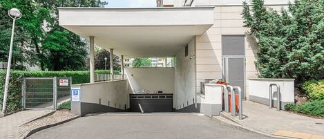 A covered entrance to a building with a gated driveway, a pedestrian pathway, and a sign indicating a parking area.