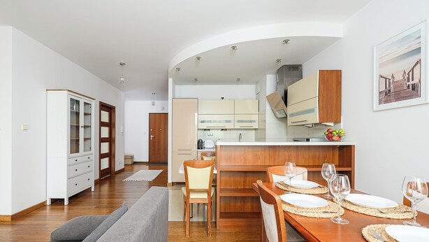 Une cuisine moderne avec des meubles en bois, un coin repas avec une table en bois dressée pour quatre personnes, et un salon avec un canapé gris.